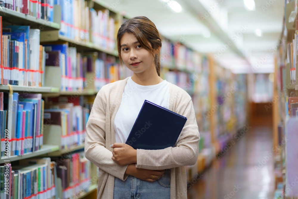 Portrait of Smart Asian woman university student reading book and looking at camera between bookshel