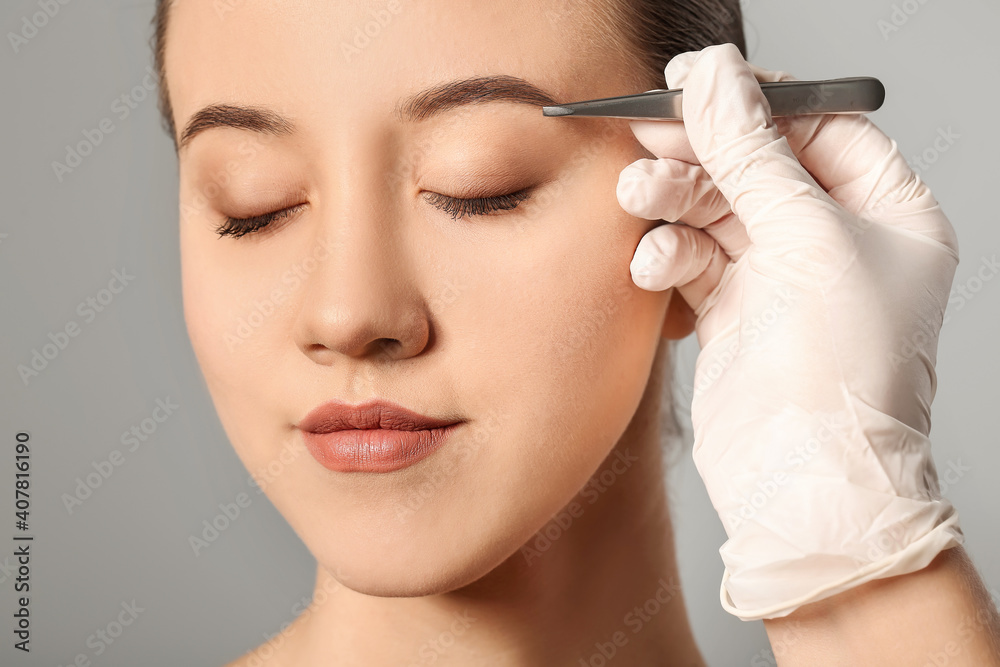 Young woman undergoing eyebrow correction procedure on grey background