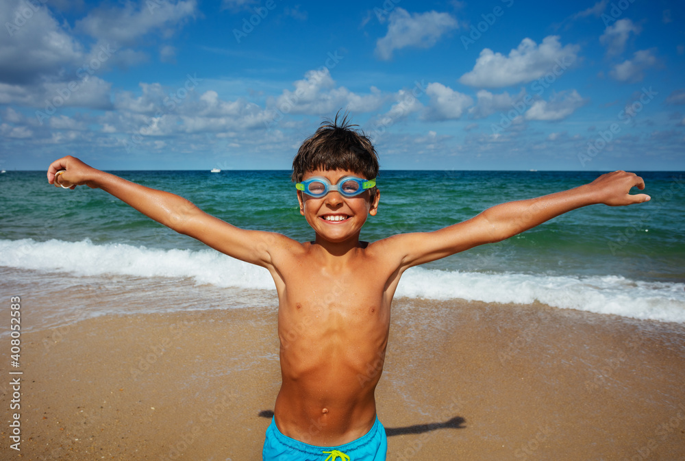 Happy smiling boy in swimming googles on the beach lift hands and smile laugh with sea on background