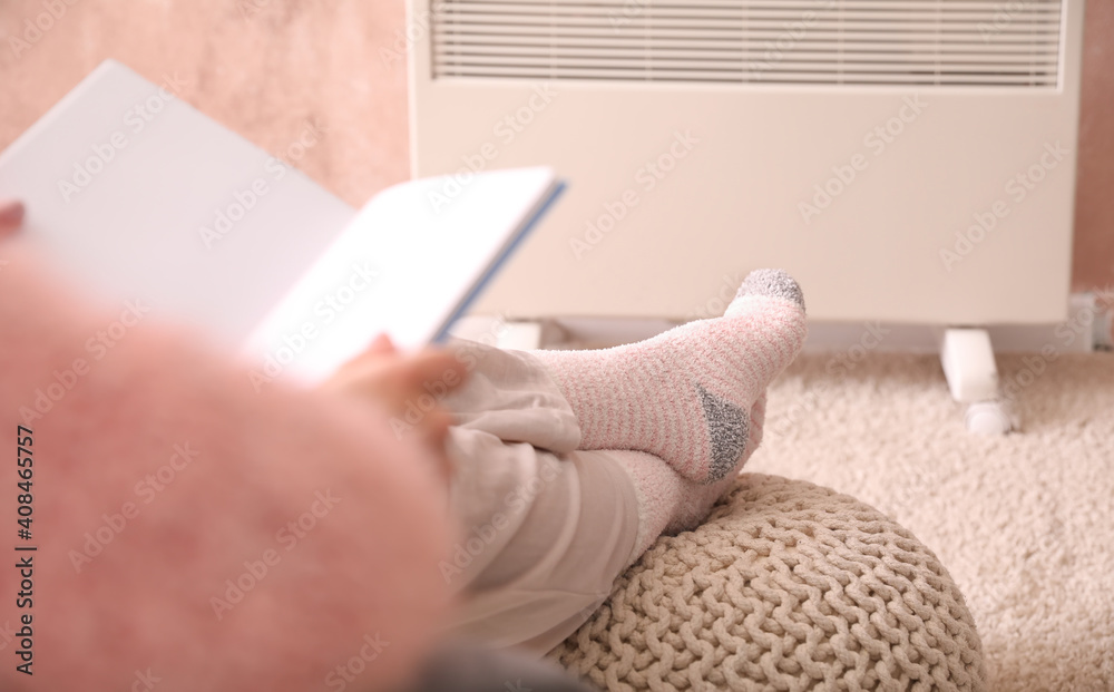 Young woman reading book near electric heater at home. Concept of heating season