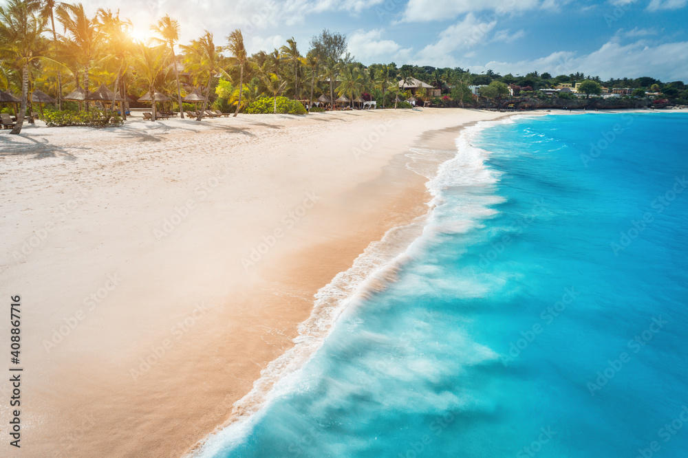 Aerial view of white sandy beach, ocean with waves, umbrellas, green palms at sunset. Summer holiday