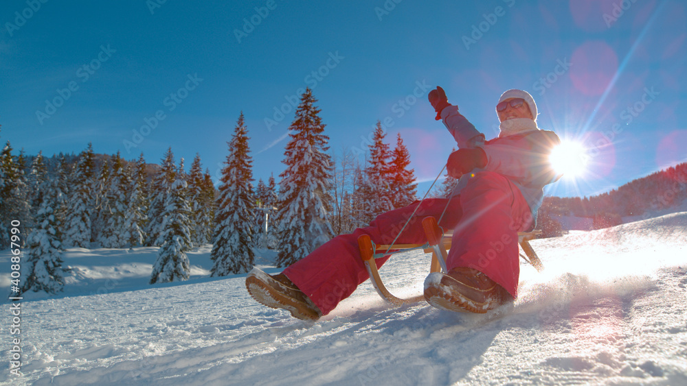 LOW ANGLE: Happy young woman rides a sled down a snowy hill on a sunny day.