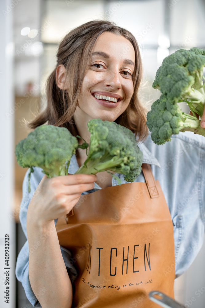 Portrait of pretty smiling woman in apron with fresh broccoli on the kitchen. Healthy vegetarian con