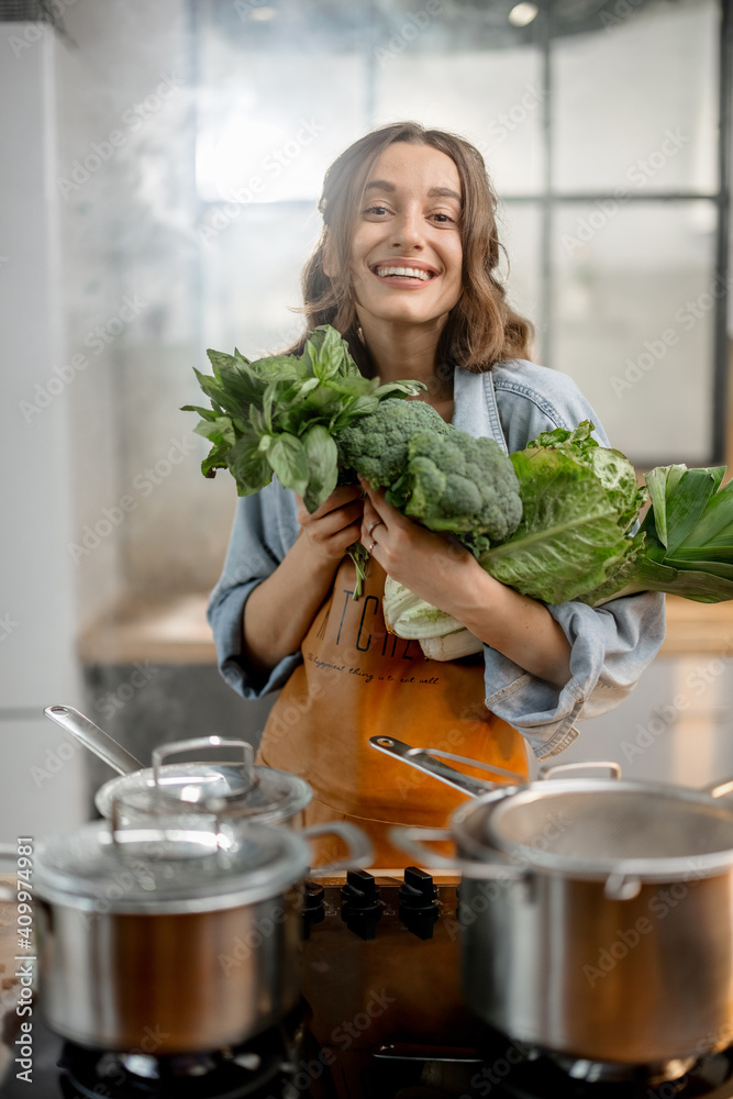 Pretty housewife in apron with fresh vegetables near cooker with boiling pans on the kitchen. Health