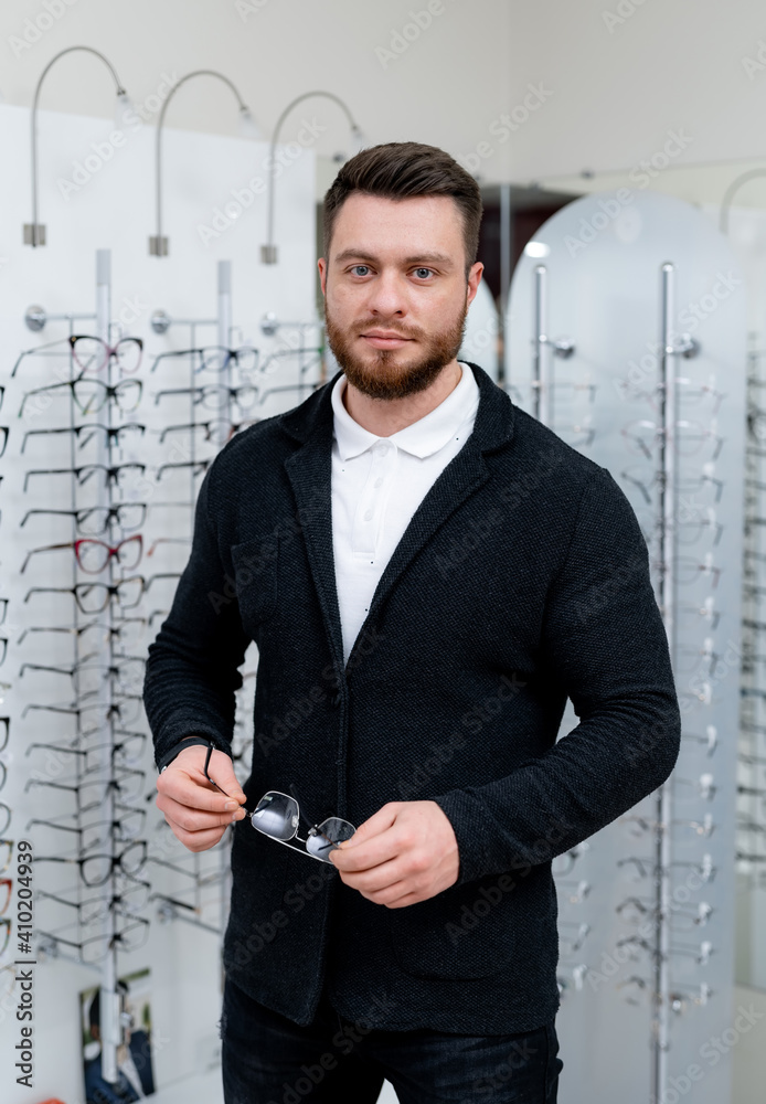 Male client is standing with set of glasses in background in optical shop. Man tries on spectacles. 