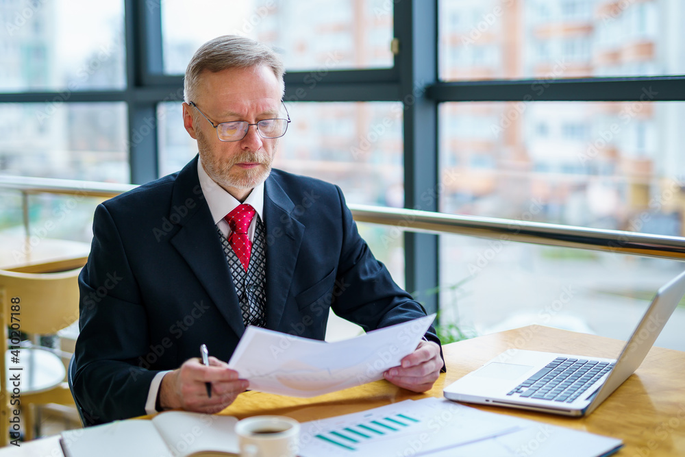 Thoughtful middle aged businessman in suit with a laptop on table while working with documents. Red 