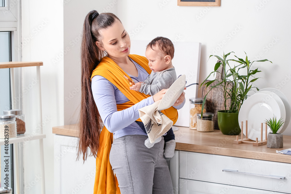 Young mother with little baby in sling washing dishes at home