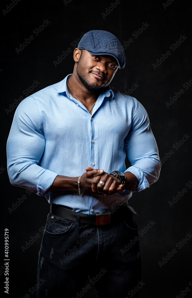 Studio portrait of black man wearing light shirt and cap posing over black artistic background. Clos