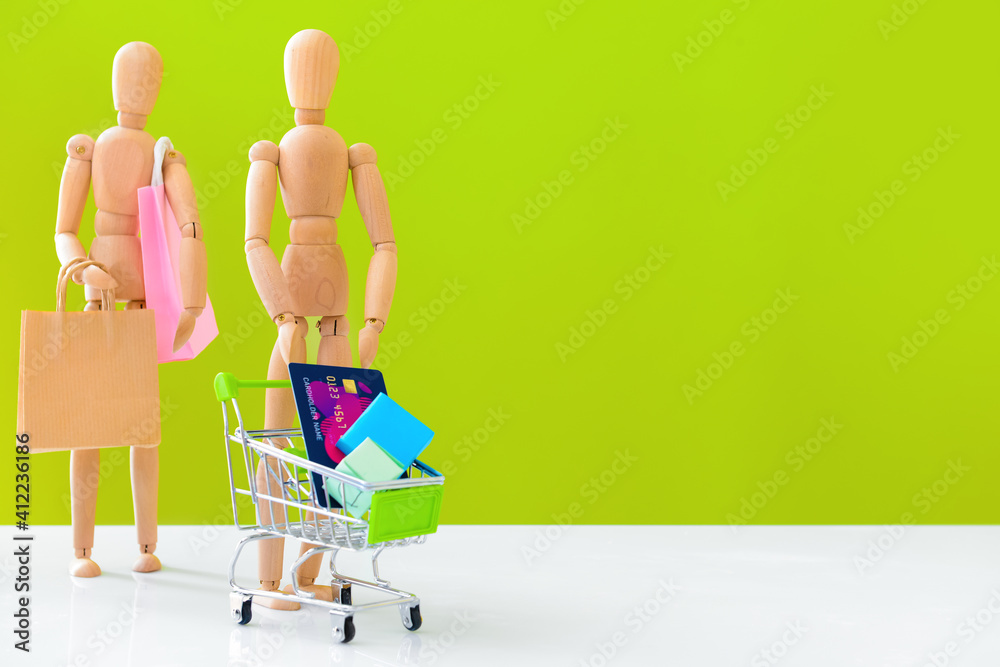 Wooden mannequins with shopping cart, bag and credit card on color background