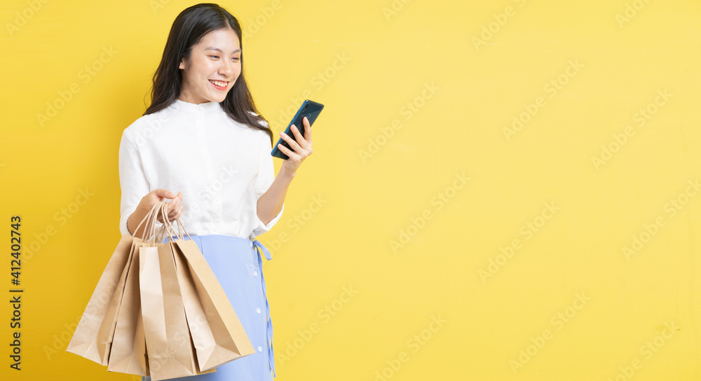 Young girl holding shopping bag with happy expression is using phone on yellow background