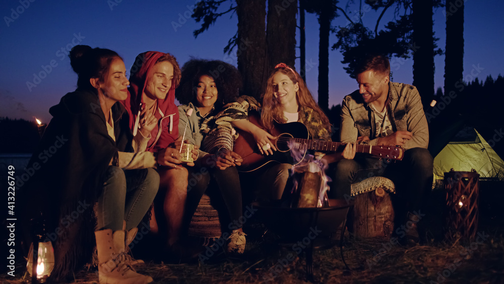 Diverse Group Of Young Men And Women Around Burning Camping Bonfire In The Woods Drinking Hot Drinks