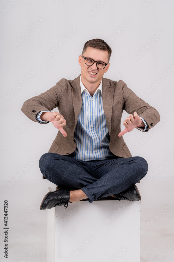 Formal man sitting on cube with legs crossed. Male showing both thumbs down, with a disguss to the c