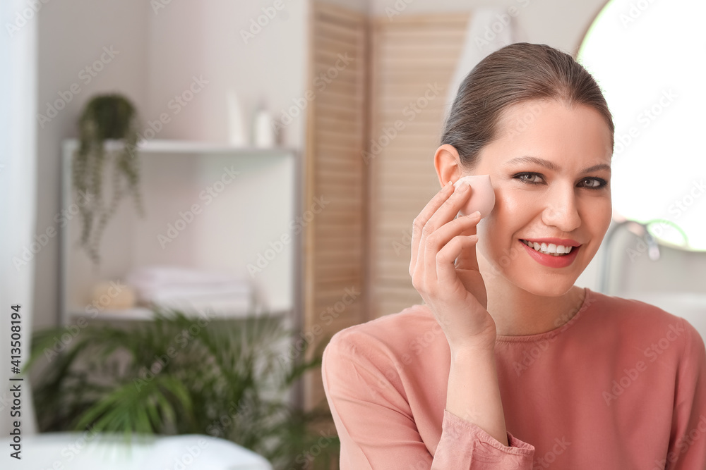 Beautiful young woman with sponge applying makeup at home