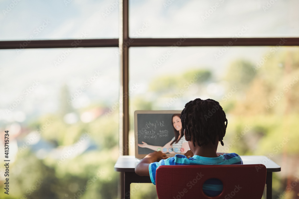 Rear view of male student having a video call with female teacher on laptop at school