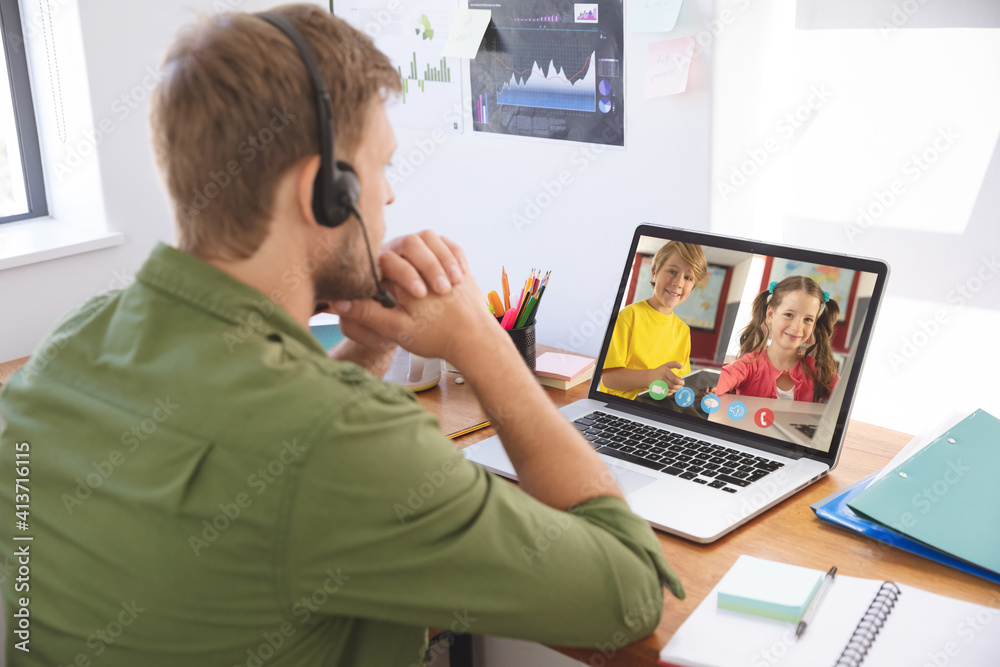 Male teacher wearing headphones having a video conference with male and female students on laptop at