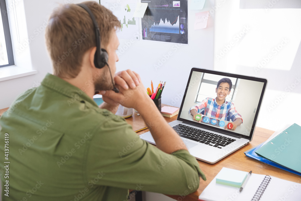 Male teacher wearing headphones having a video call with male student on laptop at school