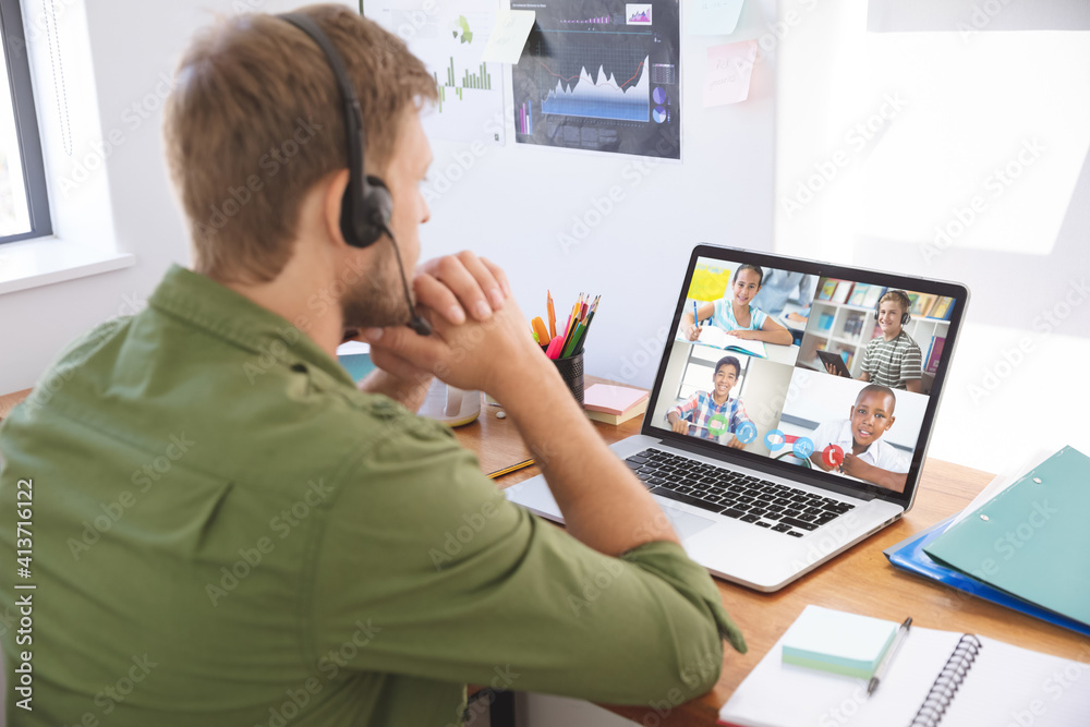 Male teacher wearing headphones having a video conference with multiple students on laptop at school