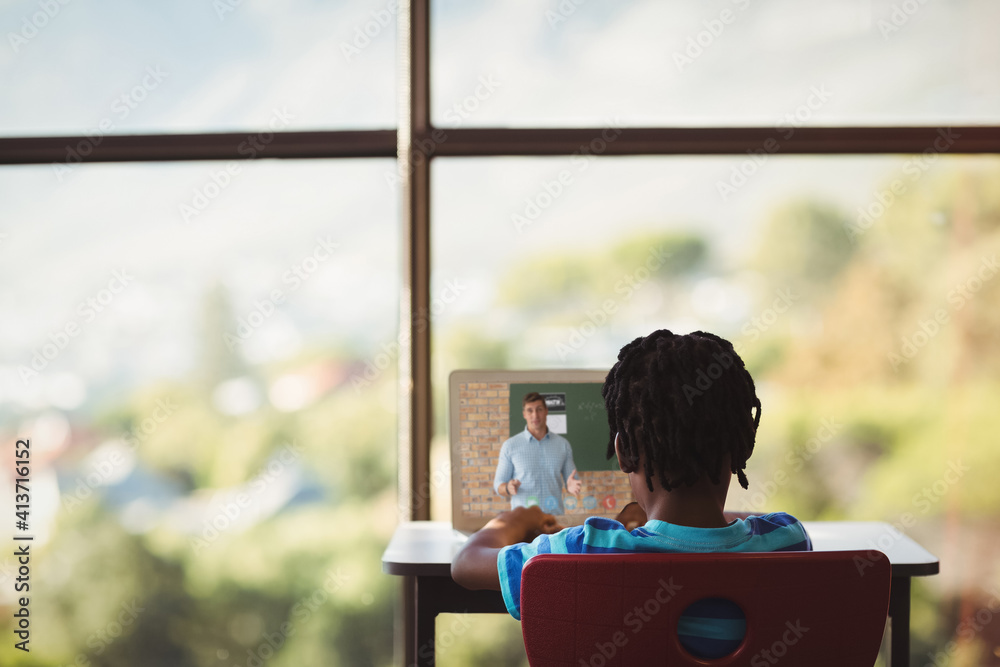 Rear view of male student having a video call with male teacher on laptop at school