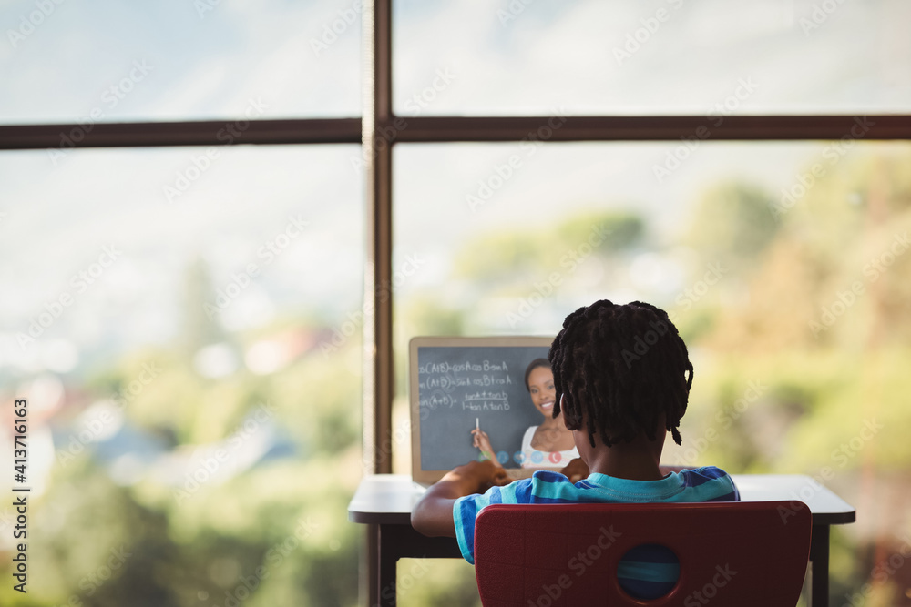 Rear view of male student having a video call with female teacher on laptop at school