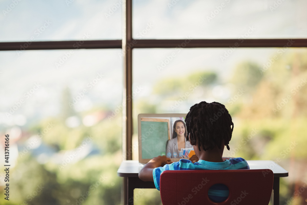 Rear view of male student having a video call with female teacher on laptop at school