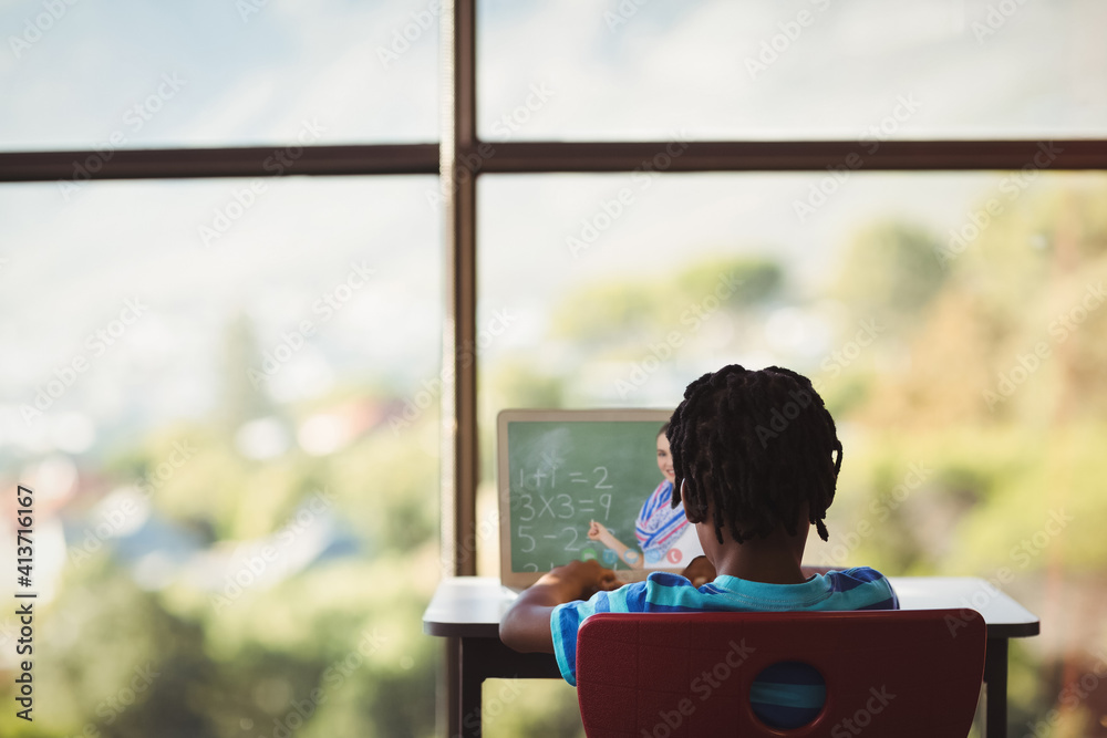 Rear view of male student having a video call with female teacher on laptop at school