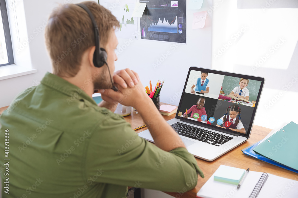 Male teacher wearing headphones having a video conference with multiple students on laptop at school