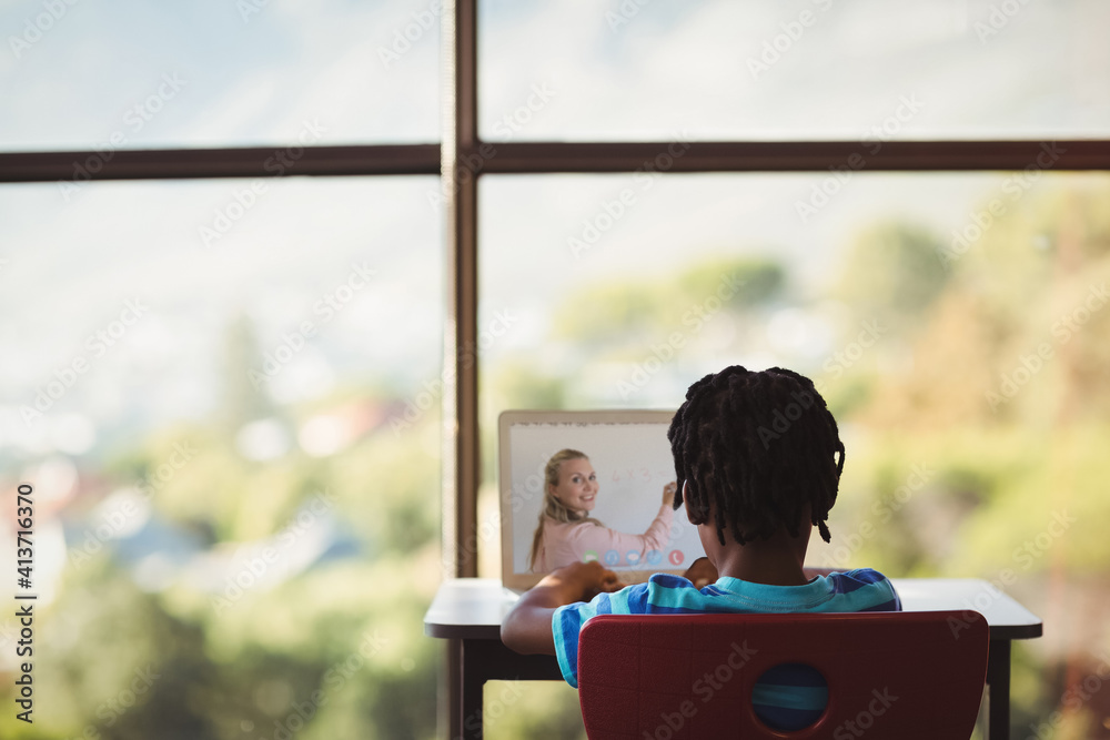 Rear view of male student having a video call with female teacher on laptop at school