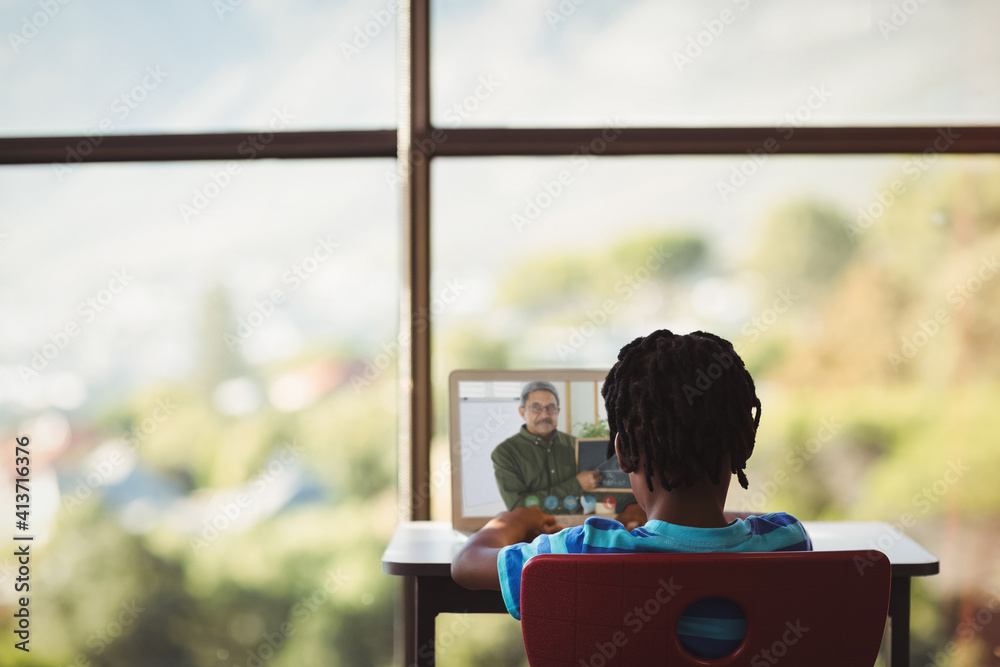 Rear view of male student having a video call with male teacher on laptop at school