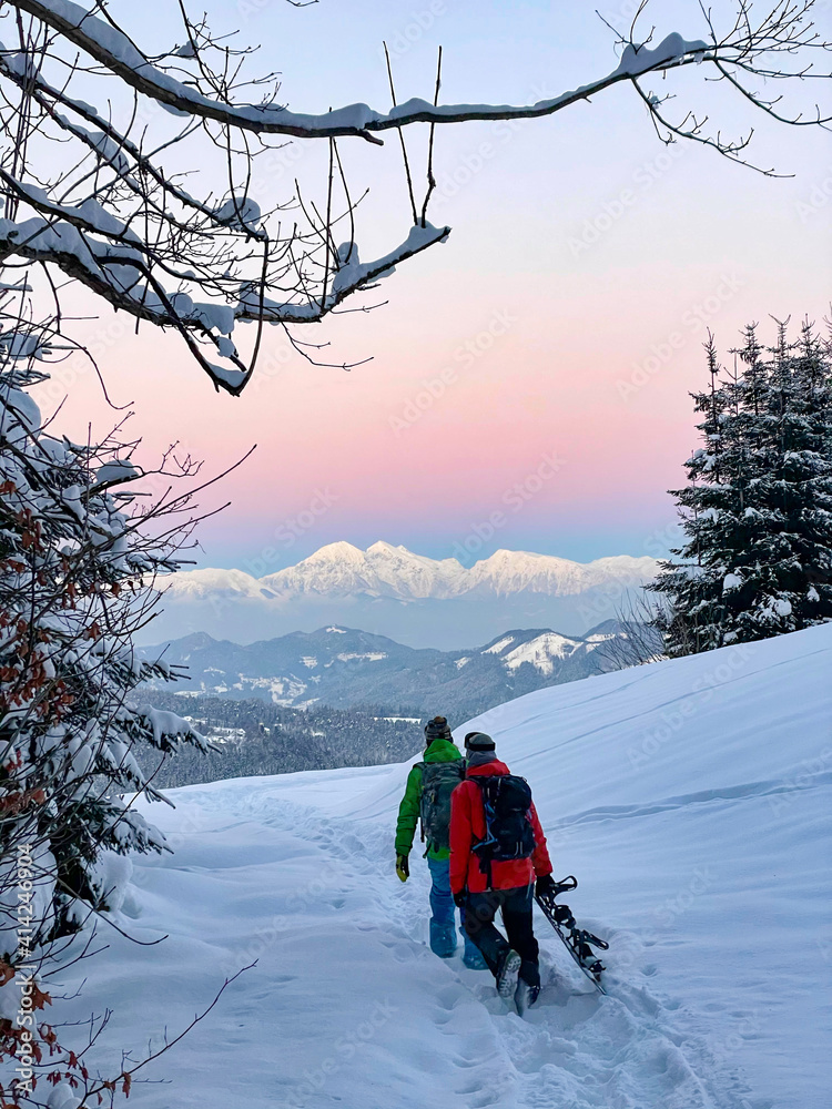 垂直：日落时，两名面目全非的单板滑雪运动员沿着雪地徒步旅行。