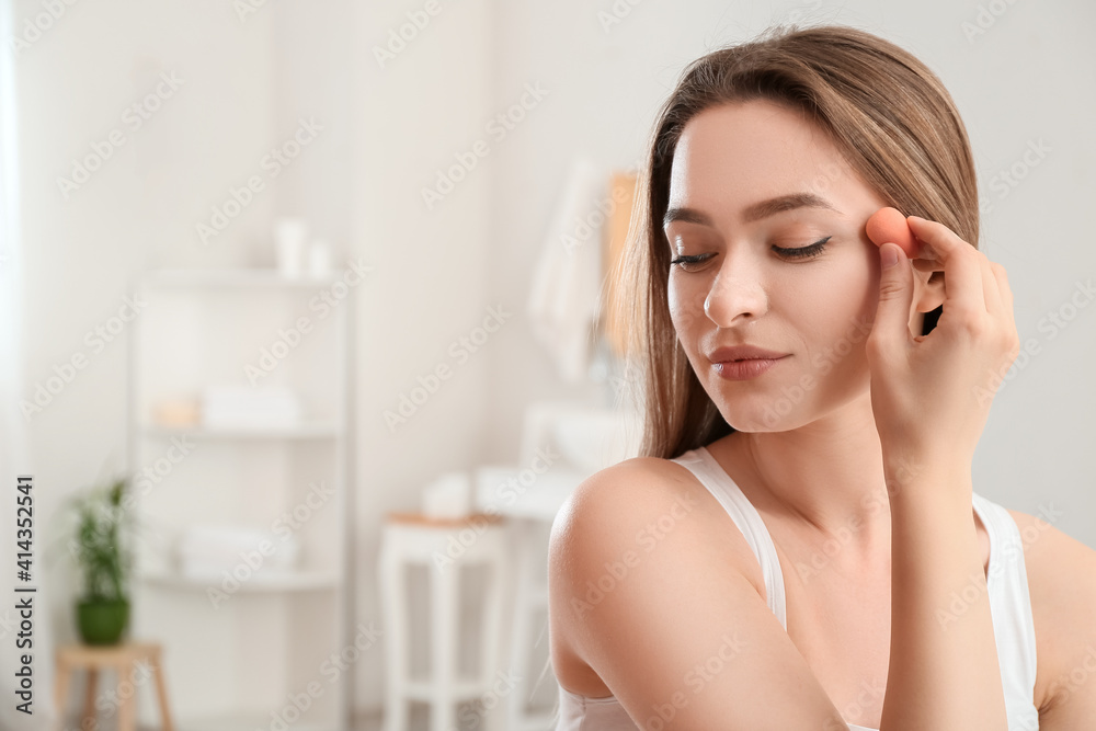 Beautiful young woman with sponge applying makeup at home