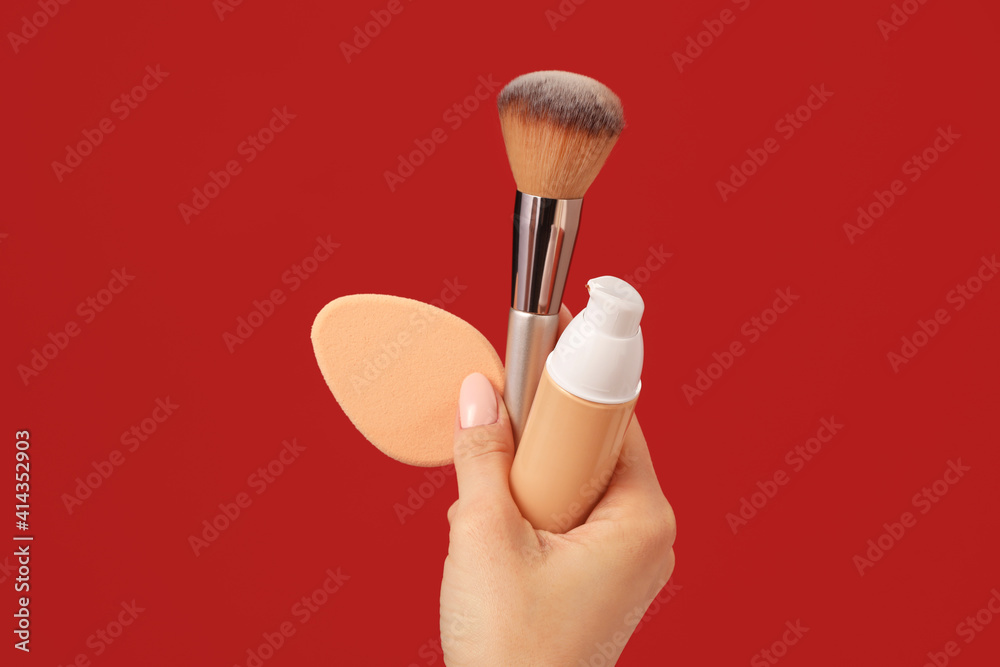 Hand with makeup sponge, concealer and brush on color background