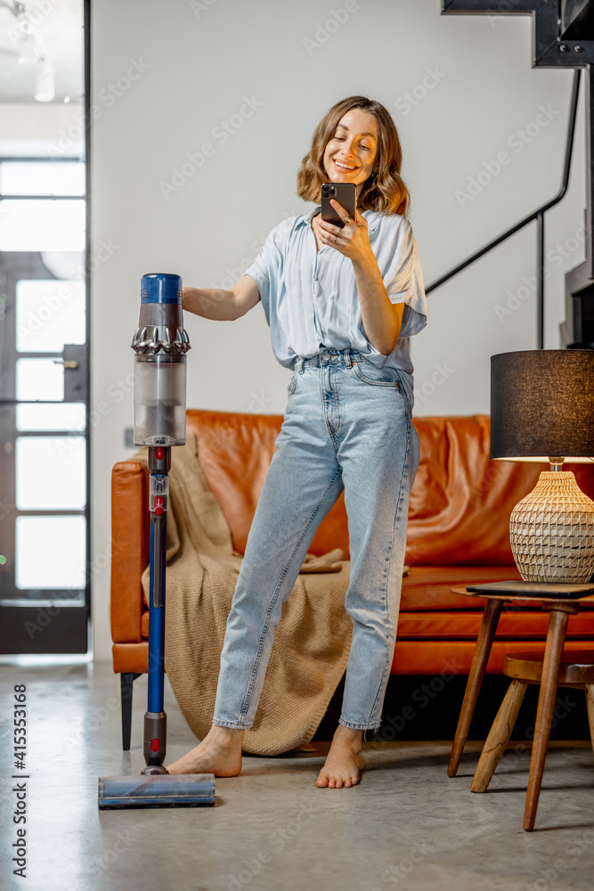 Happy woman with cordless vacuum cleaning floor and using phone at home in stylish interior design o