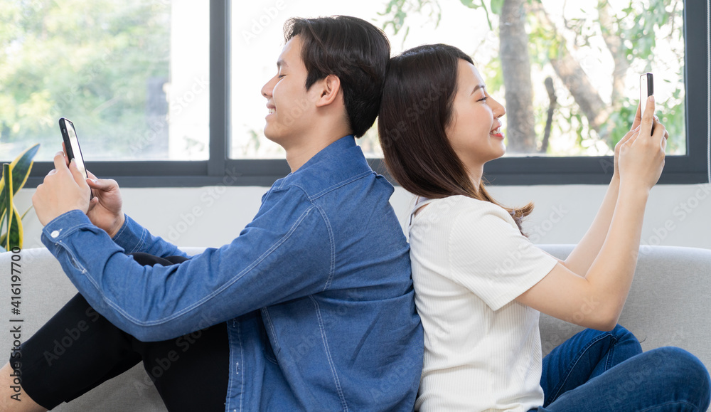 Young Asian couple sitting with their backs on each other and using the phone on the sofa