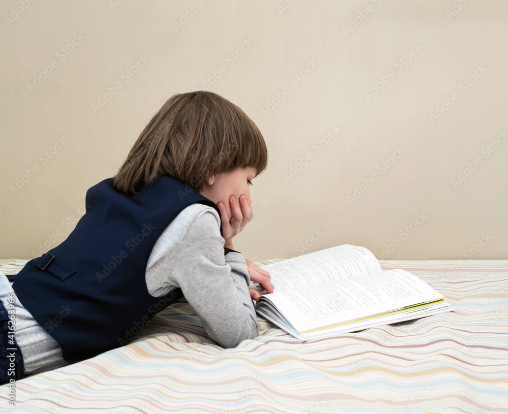 A boy in a school uniform reads a book at home while lying on the bed.