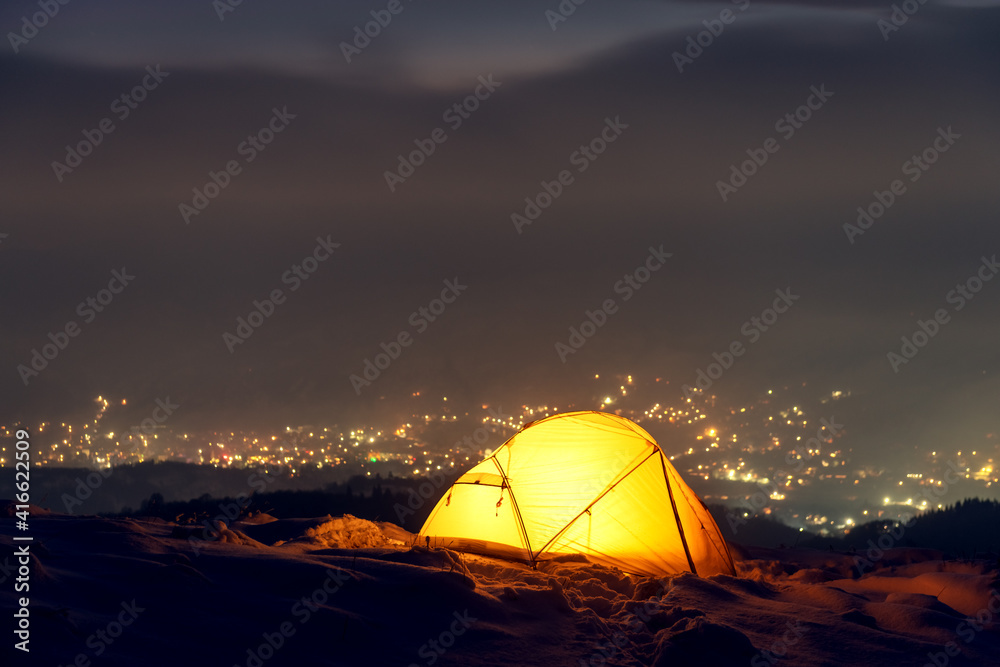 Yellow tent lighted from the inside against the backdrop of glowing city lights in fog. Amazing snow