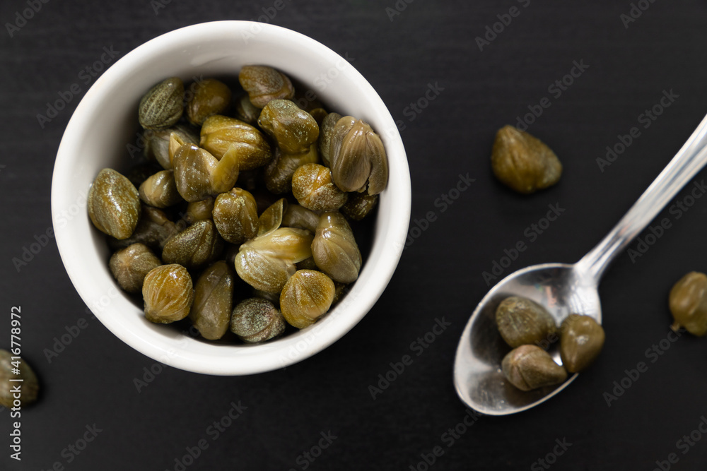 Top view of a bowl of green capers and a spoon isolated on a black background. 