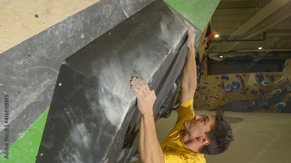 CLOSE UP: Young Caucasian male climber moves along a large black volume hold.