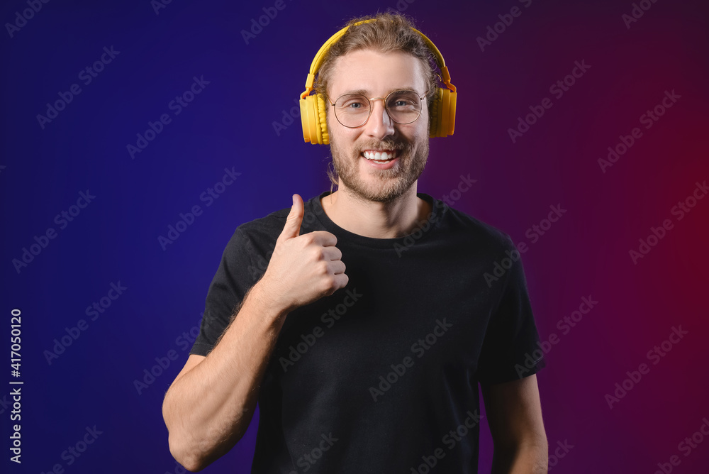 Handsome young man listening to music and showing thumb-up on dark background