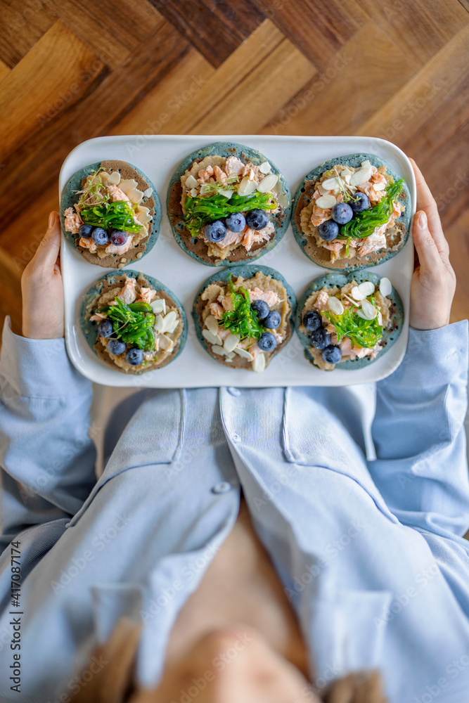 Woman in blue shirt holding a set of american pancakes on a plate with seaweed, cheese, blueberry an