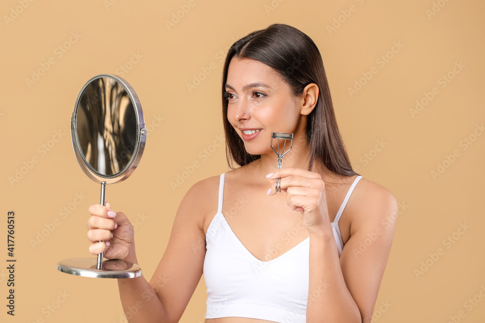 Beautiful young woman with mirror and eyelash curler on color background