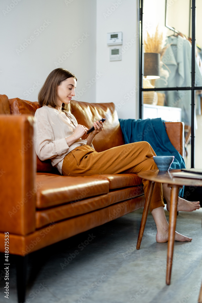 Pretty woman with Smartphone sitting on the brown sofa at home. Lifestyle and work from home concept