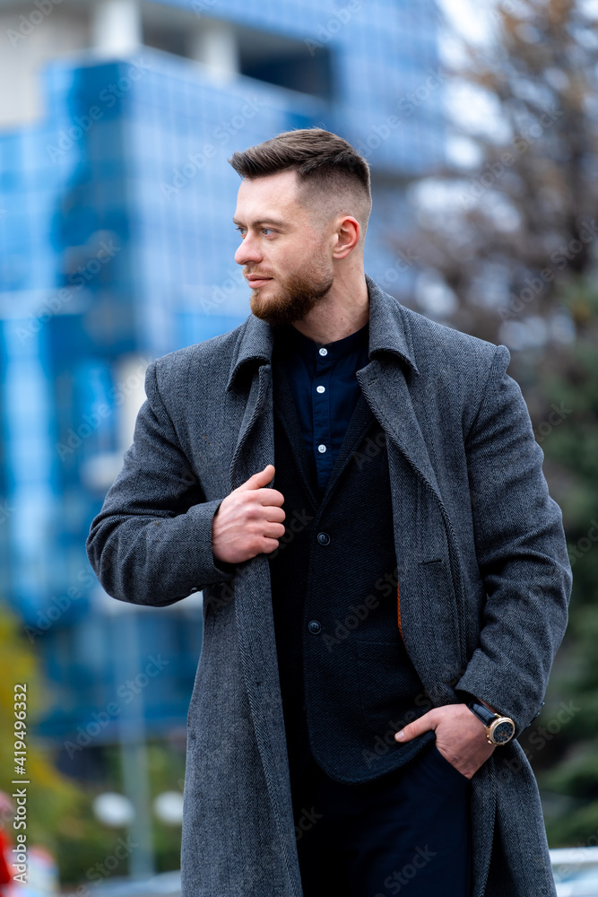 Stylish man holding hand on coat and walking near high buildings. Relaxing walk on modern street.