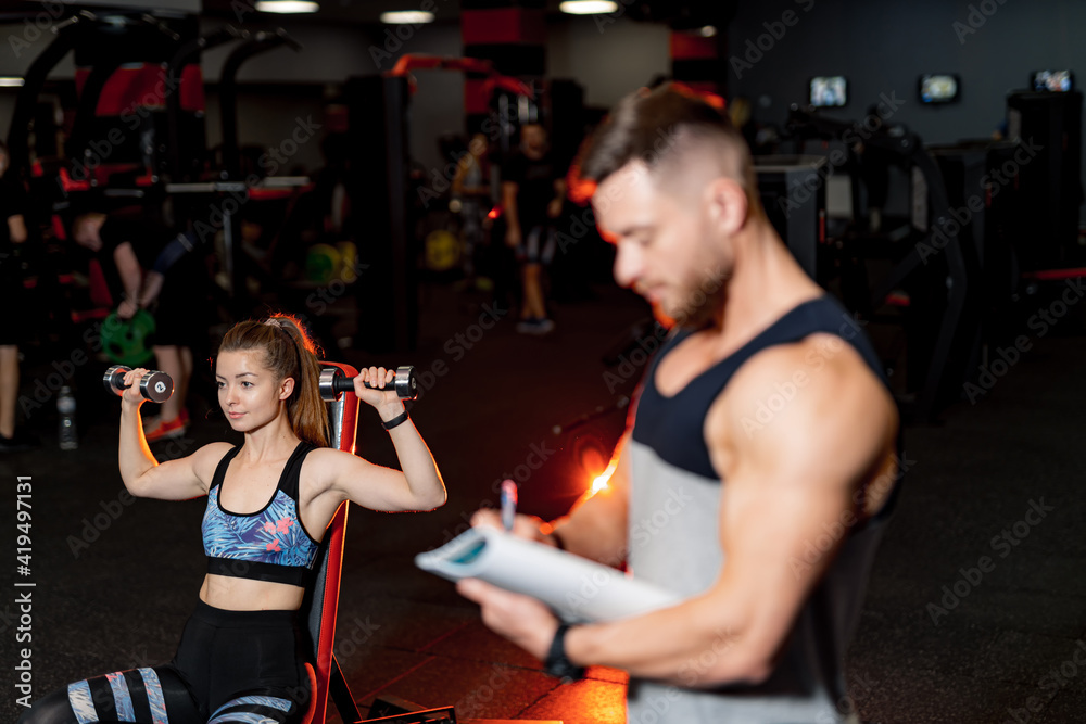 Fitness muscular trainer doing notes in clipboard while fit girl working in gym. Handsome man make u