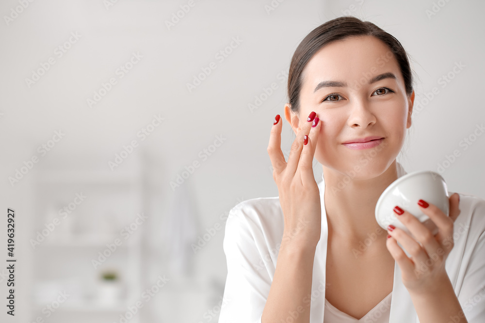 Beautiful young woman applying facial cream in bathroom, closeup