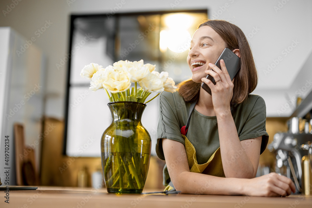 Young woman speaks on phone while staying near a bouquet of fresh tulips in stylish kitchen. Florist