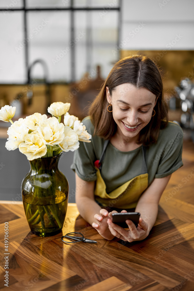 Positive young housewife using smartphone near bouquet of fresh tulips on stylish interior of kitche