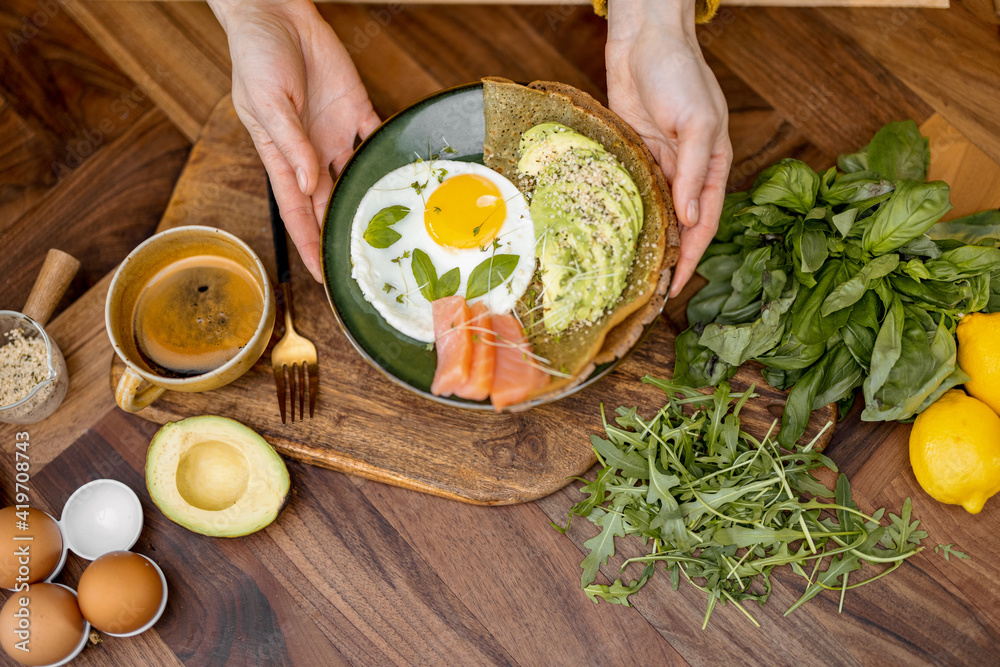 Healthy breakfast made of egg, sliced avocado, salted red fish and pancake on green plate on a woode