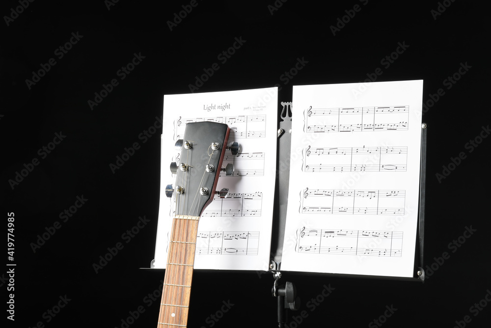 Note stand with music sheets and acoustic guitar on dark background