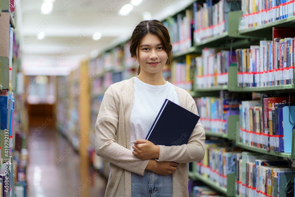 Portrait of Smart Asian woman university student reading book and looking at camera between bookshel