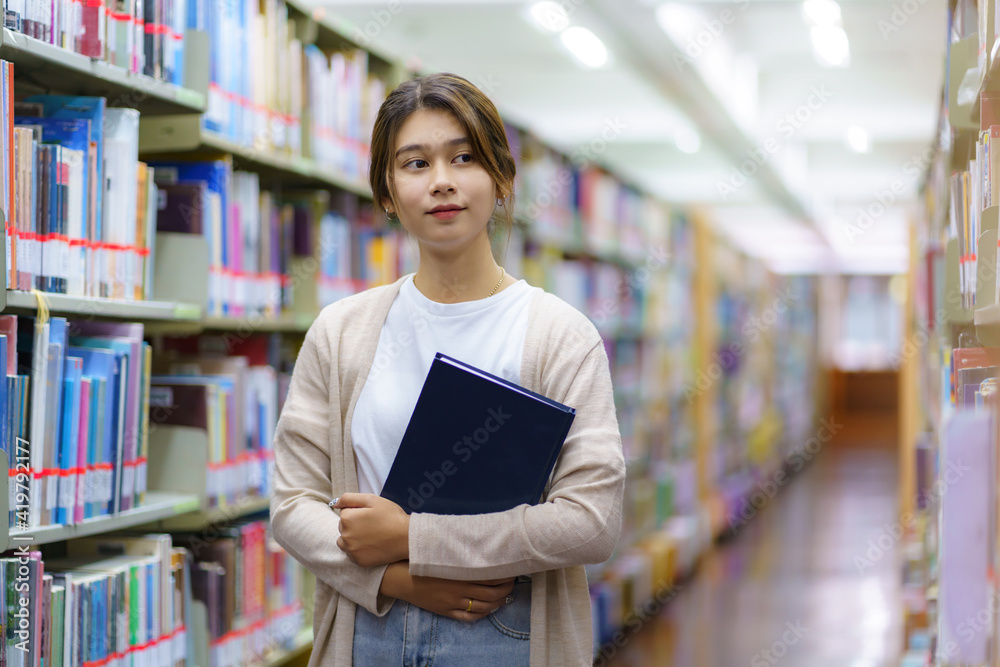 聪明的亚洲女大学生读书并看着书架之间的相机的肖像